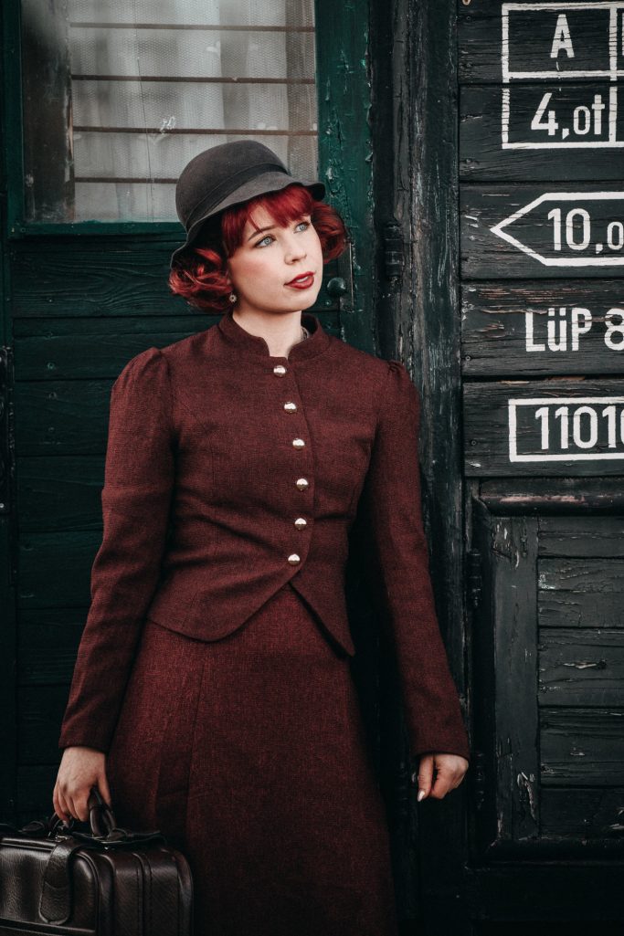 Frau mit rotem Haar in einem braunen Kleid und Hut, hält einen Koffer.