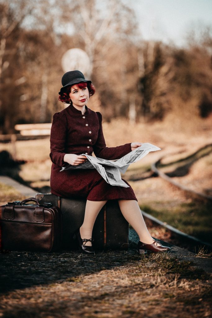 Frau in vintage Kleidung sitzt auf einem Koffer und liest eine Zeitung im Freien.